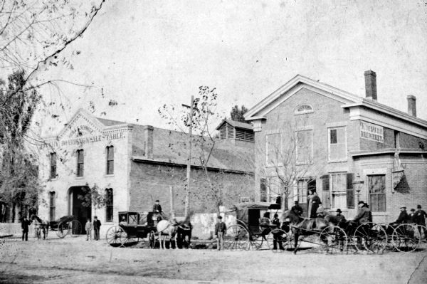View across State Street towards two buildings, with, on the left, the Hess & Schmitz Livery Boarding and Sale Stable, and on the right the Empire Brewery. There are horse-drawn vehicles outside both establishments. Three women are standing in the doorway of the brewery.
