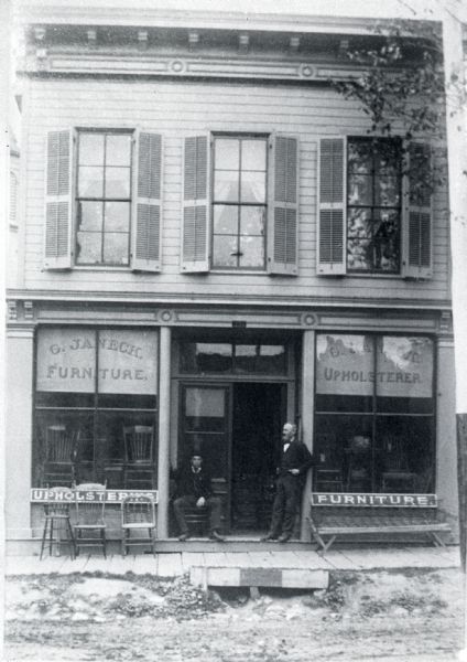 View across unpaved street towards two men wearing suits, vests and ties posing in the doorway of the C. Janeck Furniture and Upholstery store on State Street. In front of the left display window are three wooden chairs on the board sidewalk, and inside the window more chairs are on display. On the right is a settee or bench, and more chairs on on display inside the window.