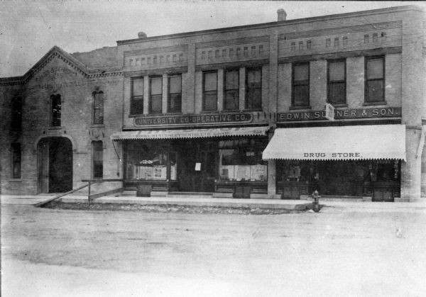 View across street towards the University Cooperative storefront, which had multiple locations over time. This one is at 504-506 State Street. The awning reads: "Books, Stationary, Athletic Goods." Next door to the right is the "Edwin Sumner and Son Drug Store." On the left is the Hess & Schmitz building. There is a fire hydrant on the curb in front of the drug store.