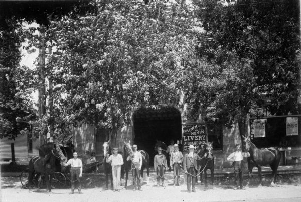 View across street towards people posing in front of the Brown and Nevin Livery, Boarding and Stables at 508 State Street. There are trees along the sidewalk. A number of the men are holding horses by leads or halters.