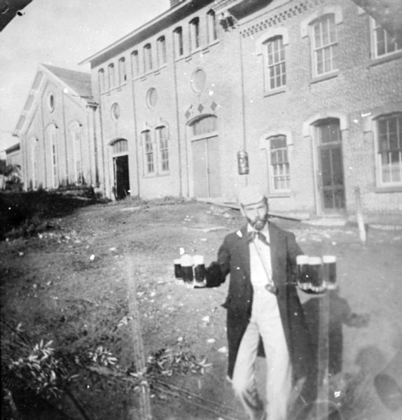 A man is posing outdoors in front of the Hausmann Brewing Company Malt House at 1603 Sherman Avenue. He is standing and smoking a cigar and carrying three mugs of beer in each hand.