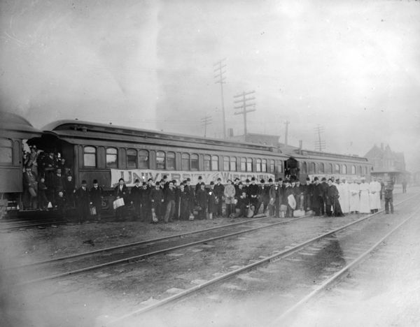 View across railroad tracks towards a group of glee, banjo and mandolin club members posing in front of a University of Wisconsin banner on the Chicago and Northwestern train. More people are posing in the doorways of the train cars. On the far right are railroad employees posing with the group. The Chicago and Northwestern station was at 620 E. Wilson Street.