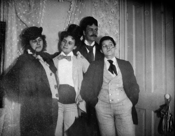 Four women dressed as men posing togther for a portrait. The woman on the left is wearing a beard and has a pipe in her mouth. In the center is a woman wearing a striped suit, a cummerbund and a bow tie with a top hat. Third from the left is a tall woman wearing a mustache and a necktie. On the far right is a woman smoking a cigarette and wearing a coat over checked pants and matching vest. They are standing in a room near a door in front of a window with dotted curtains.