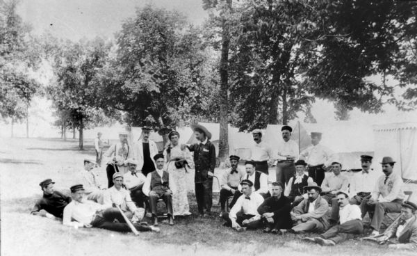 Outdoor group portrait of Western Canoe Association members lounging in front of their tent camp. They are all wearing caps or hats. Near the center of the back row, one of them is wearing a cap and a kimono or bathrobe and holding a catcher's mitt. A man next to him appears to be wearing pajamas. On the ground in the front row the second man from the left is holding a baseball bat. Stanley C. Hanks was a member of this group. In a canoe sailing race to Picnic Point on July 13, 1894 for the Gardner and Longworth Cup Heats, Stanley Hanks won second place.