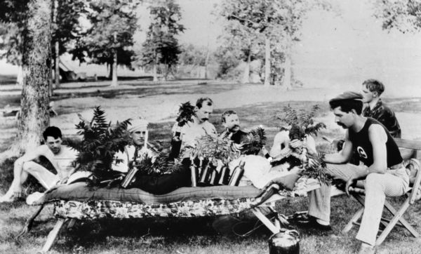 Seven members of the canoe association are posed around an eighth member, who is lying face down on a padded cot, covered with ferns. Ferns have been strategically placed, including one man holding them in front of his face. On the left in the background is a tent at Picnic Point.
