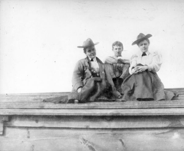 View looking up at a man sitting between two women on a roof. The women are wearing hats, skirts and collared blouses, and the woman on the right is wearing a blouse with leg o'mutton sleeves. The woman on the left is wearing a jacket.
