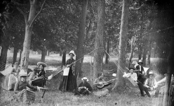 A group of people gathered on and around hammocks in a wooded area. Three women are sitting in a hammock on the left, and on the right three boys are sitting in a hammock. Behind the boys is another hammock with two boys. In the center is a woman standing, a man whittling, and a girl playing a guitar, and in the left foreground is a woman sitting in the grass leaning on a camp stool.