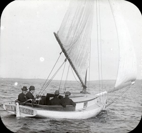 View across water towards four men wearing suits and hats sitting in a sailboat.