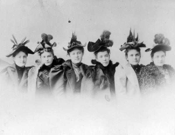 Vignetted group portrait of six young women posing in a row. They are all wearing hats with flat crowns, curved or bent brims and lavish feather arrangements. They are also wearing winter coats with large, puffy sleeves. Two women have fur trim on the collars.