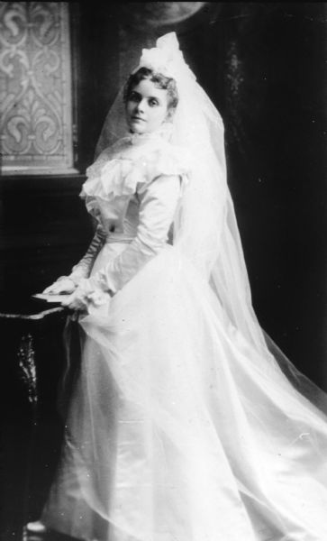 Full-length portrait of Ina Judge wearing her wedding dress while standing in front of a painted backdrop or in a church. Ina Judge married Stanley Hanks on December 1, 1897.