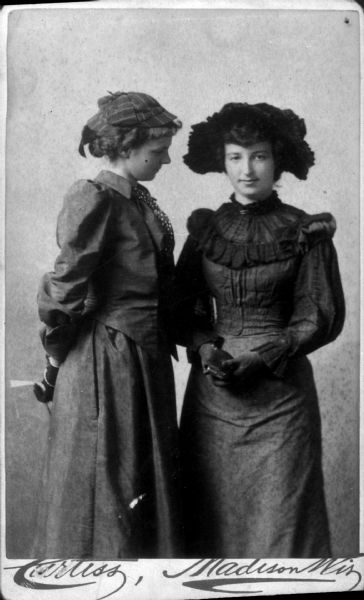Three-quarter length portrait of two women standing and wearing dark dresses and hats. The woman on the left stands sideways, looking at the other woman, and has her hands behind her back holding what may be a hand fan. The woman on the right is facing forward and holding something in her gloved hands.