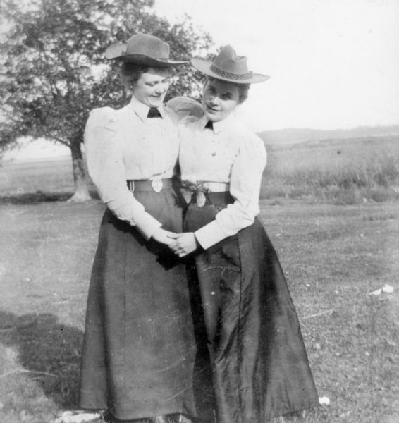 Full-length portrait of two women standing on a lawn, holding hands and smiling. They are wearing cowboy hats, white shirt waist blouses with puffy sleeves, and long dark skirts with matching belts.