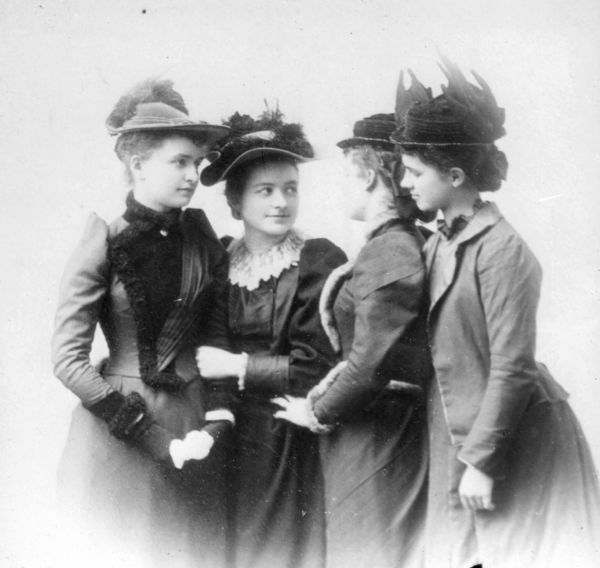 Vignetted group portrait of four women standing closely in a row gazing at each other. The two women on the right stand in profile looking at the other two women. The women are wearing tight-fitting jackets with puffy sleeves over long skirts, and elaborate hats with small brims, low crowns and feathers.