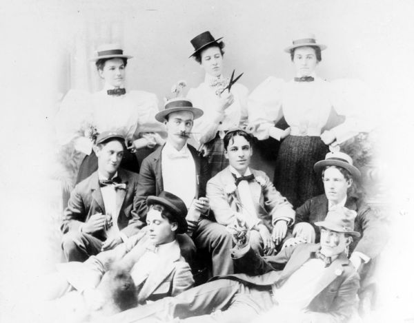 Studio group portrait of nine people. Three women are standing in the back row, and the woman in the middle us holding a pair of shears or scissors. The three women are wearing hats, white blouses with puffy sleeves, and neckties, skirts and wide belts at their small waists. The six men in the front are sitting or lounging in casual poses, and some are holding cigarettes. They are wearing suits, hats or caps, and bow ties.