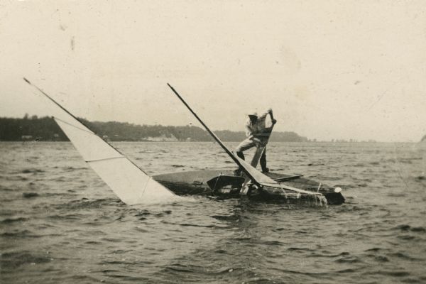 View across water towards a man working to turn a sail canoe, rolled over onto its left side, upright. The canoe has two sails, is dark-colored, and has a single seat. The man appears to be using the tiller for leverage. In the background is the far shoreline.