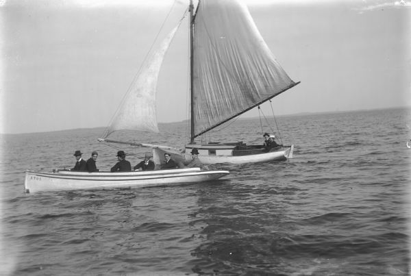 View across water towards two boats close together on a lake. There are six men in the boat in front, one man with a pipe in his mouth, and two men in the sailboat behind it. In the background is the far shoreline.