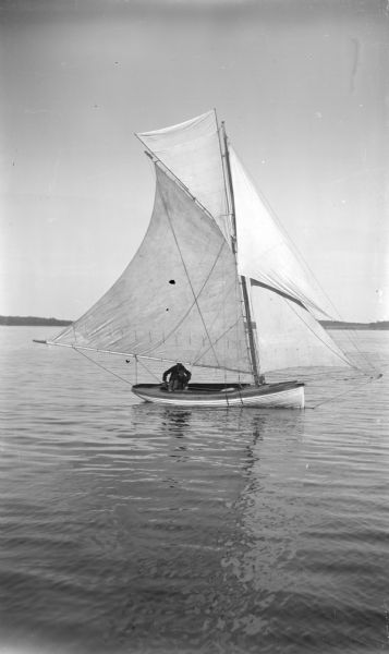 View across water towards two men in a small sailboat with large sails.