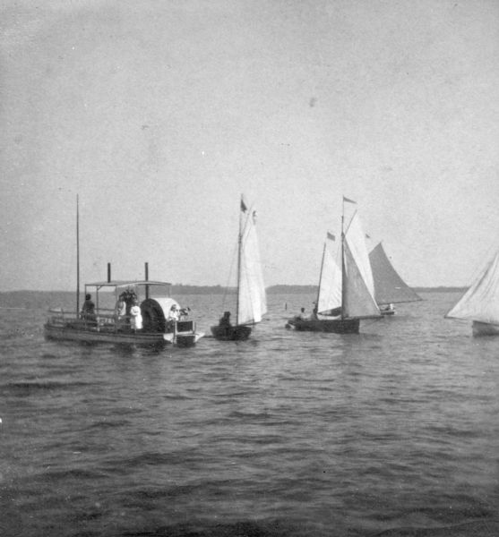 View across water towards women on a steam paddle boat on the left, and four sailboats near them on the right.