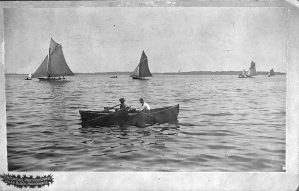 View across water towards two men in a rowboat who are looking out at sailboats on Lake Monona. In the background is the far shoreline.