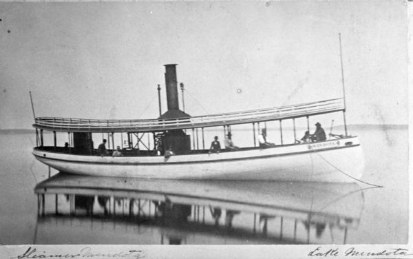View across water towards the steamboat "Mendota" reflected in the water of Lake Mendota with at least nine people on board. The far shoreline is in the background.