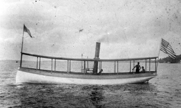 View across water towards the steamboat "Satirio," owned by Henry Ainsworth. There is an American flag flying from the prow, and a banner hanging off the end. There are three people on board.