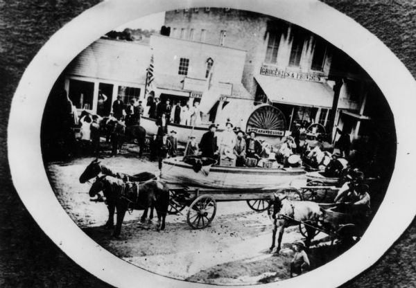 Elevated view of the steamer "Scutanawbequon" being carried by a wagon to the lake for launching. This is probably a street on the Capitol Square. Many people are watching, and groups of people are in boats in other horse-drawn wagons. Behind the steam drum with the name "Scutanawbequon" on it is a store with a sign that reads: "Groceries and Provisions." The steamboat was launched on April 17, 1866.