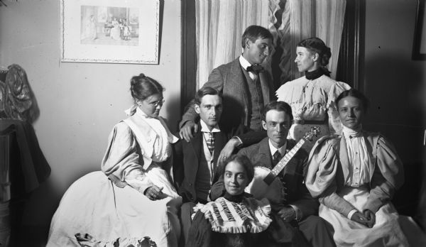 Group portrait of four women and three men posing together in a room in front of a window. The man in the center is holding a banjo in his lap. The men are wearing suits and neckties, and the woman are wearing dresses with puffy sleeves.
