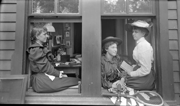 Outdoor view of three young women sitting windowsills of two open windows. The woman on the left is sitting in profile facing the two women in the windowsill on the right. One woman on the right is leaning through the window and holding what may be a firecracker. On the windowsill is a tennis racket on an American Flag. The woman on the far right is sitting and holding a book ("Better Dead My Lady Nicotine, by J.M. Barrie"). In the room behind the women is a desk with a lamp and a vase of flowers.