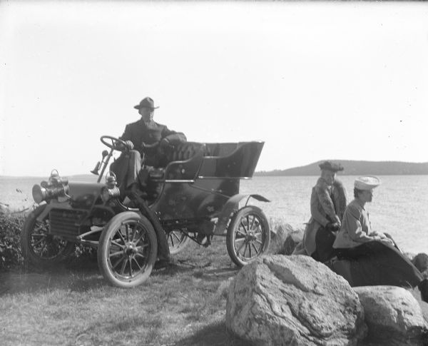 View of A.G. Zimmerman sitting in his automobile near the lake shore. He has one arm resting on the steering wheel; the other on the seat back. Two women are seated on rocks overlooking the lake in the background. The far shoreline is the background.