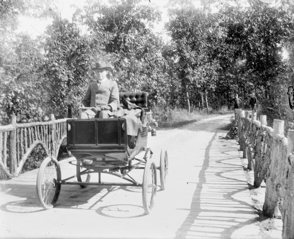 View looking towards A.G. Zimmerman driving a horseless carriage, or early automobile over a rustic bridge. The bridge has board flooring and railings made of bent saplings. Albert is wearing a suit, hat, tie and leather gloves. Two men are standing on the unpaved road in the background.

This is the Shorewood Hills rustic bridge, a picturesque highlight of Lake Mendota Drive. The Park and Pleasure Drive created the drive a decade earlier for scenic excursions in horse-drawn vehicles. To prevent trouble with the horses, early autos and carriages used the pleasure drive on different days.