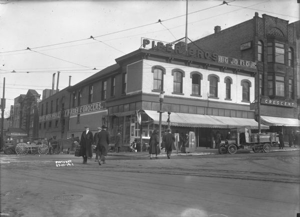 View across intersection towards the Piper Brothers Bakers and Grocers shop on the corner of Pinckney and Mifflin Streets. Other stores on that block, from the left, are Printing, Capitol Alleys and Billiards, and the Madison Federation of Labor. The Piper Brothers shop has a large electric sign advertising "Big Jo Flour." In front of Piper's is a "News Stand" on the sidewalk, and a large pole with an elevated sign for the streetcar that reads: "Wingra Park, West Main, Johnson St. Cars Stop Here." A storefront on the right reads: "Crescent." Numerous men and women are walking on the sidewalks and crossing the streets. There are trucks and horse-drawn vehicles parked at the curbs.