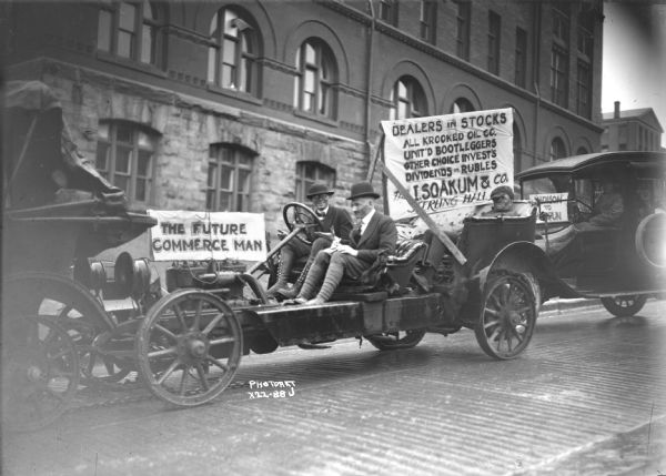 Three students, apparently in costume, are riding in an automobile outside the University of Wisconsin Armory or Red Gym with a sign over the exposed engine that reads: "The Future Commerce Man." The vehicle on the far left appears to be pulling this car with a chain, and the man sitting in the driver's seat is holding his hands away from the steering wheel. Another sign next to the man in the backseat reads: "Dealers in Stocks, All Krooked Oil Co., Unit'd Bootleggers, Other Choice Invest's, Dividends in Rubles. The I. Soakum & Co., Sterling Hall." The automobile on the far right has a sign offering Madison to Waupun rides.