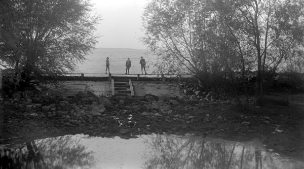 View across pond towards three boys standing on the board walkway of the spillway at Lake Mendota and the Yahara River. In the background is the far shoreline.