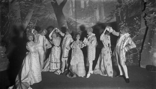 Slightly elevated view of four couples, dressed in late eighteenth century costumes, on the stage of the Fuller Opera House. They are posing as if they are dancing. Behind them is a painted backdrop.