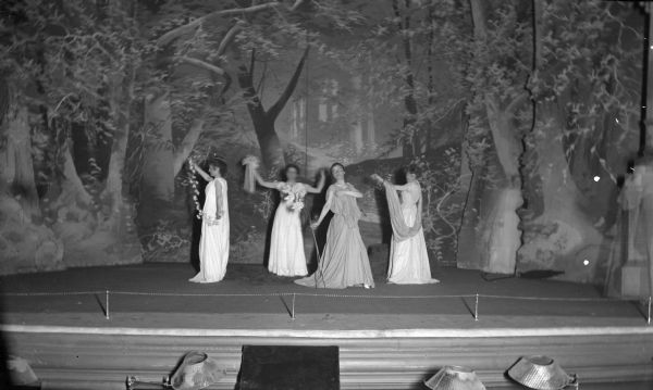Slightly elevated view of four women in flowing, classical dresses dancing on the stage of the Fuller Opera House. Behind them is a painted backdrop.