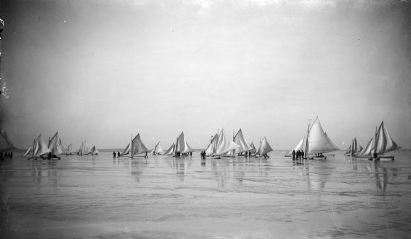 View across ice towards the crews of ice yachts standing near their iceboats. There are seventeen or eighteen iceboats, and their are numbers on the sails.