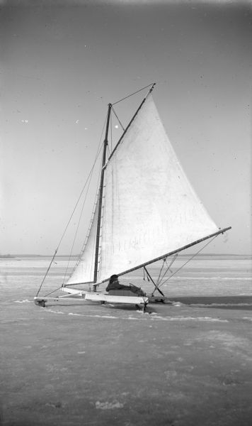 View across ice towards two people in an iceboat on Lake Mendota.