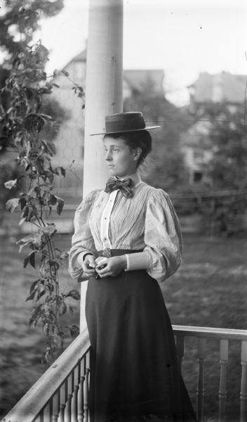 Three-quarter view of a woman dressed in a dark summer skirt and a striped blouse with leg-of-mutton sleeves and a striped tie, standing on a porch looking off to the left. Her hair is up and she is wearing a straw boater. She is holding a handkerchief in her hands.