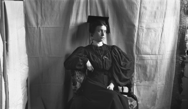 A young woman is posing in an upholstered chair in front of a cloth backdrop for her graduation portrait. She is wearing a mortar board with a tassel, a dark dress with leg-of-mutton sleeves, and a dotted collar with a large bow in the back.