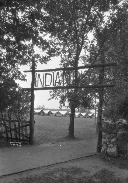 View of the entrance to Camp Indianola on the north shore of Lake Mendota, which was one of the nation's oldest camps. In the background across the lawn is a row of tents. Over 20 buildings once stood on the campgrounds. The site is now part of Governor Nelson State Park.