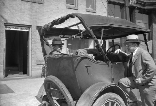 Outdoor view of a man, probably Stanley Hanks, smiling broadly and sitting in the backseat of a dilapidated car. He has one leg up on the back of the front seat. On the right, standing at the side of the car is a man standing with one leg on the running board and an arm on the front seat. In the background is an open doorway to the Photoart House.