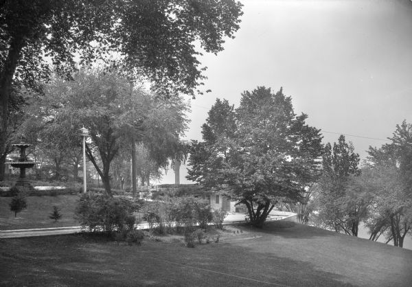 Elevated view towards the right side of the grounds and Capitol Fountain of the Executive Mansion, which was located at 130 East Gilman Street. There is a birdhouse on a pole near the fountain on the left, and a driveway along the side of the property leading down to the garage. Lake Mendota is in the background on the right.