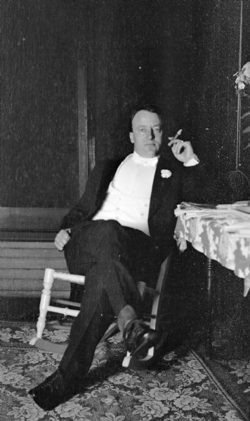 View of a man sitting in a wooden rocking chair on the porch of the Hanks home smoking a cigar.