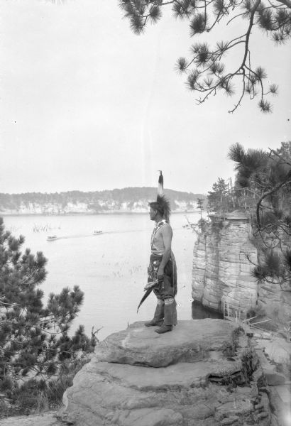 A Native American Indian in native dress, including a beaded bandolier and feathered headdress, gazing out to the Wisconsin River. He is standing on a rock high above the river, and two excursion boats are coming down the river. There is a wooden boat landing at the base of the cliff in the background on the right. A group of Native Americans, including Ho-Chunk, Sioux, Kiowa and southwestern Native American tribes, were gathered at Wisconsin Dells for the Stand Rock Indian Ceremonial.