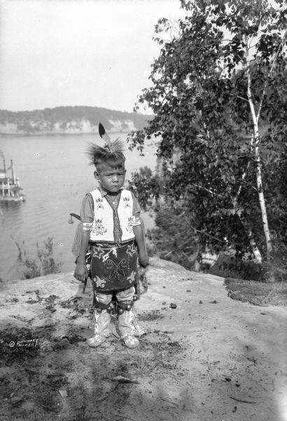 A young Native American Indian boy in traditional dress is posing on a rock formation above the Wisconsin River. In the background is a steamboat on the river. The boy is wearing a feathered headdress, embroidered or beaded vest and apron. Around his knees are bell bands,  and he is wearing beaded leather boots. A group of Native Americans, including Ho-Chunk, Sioux, Kiowa and southwestern Native American tribes were gathered at Wisconsin Dells for the Stand Rock Indian Ceremonial.