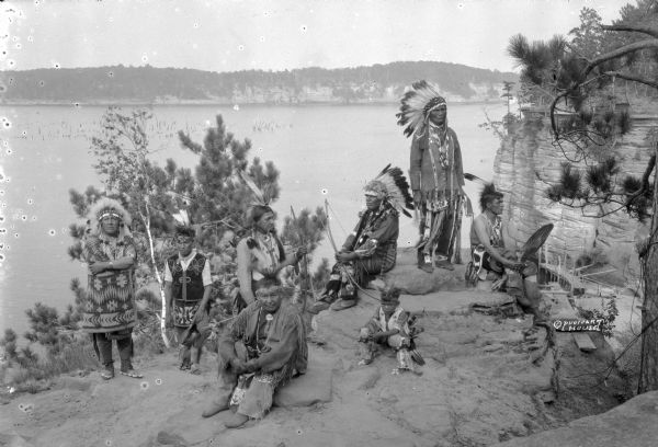 Group portrait of older men, with younger men and a boy, posing on a rock formation above the Wisconsin River. They are all wearing traditional clothing, including feathered headdresses, beaded or embroidered shirts or vests, beaded moccasins or boots, and a variety of necklaces. Several of them are holding weapons. Others are wearing bell bands on their legs. There is a wooden boat landing at the base of the cliff in the background on the right.
A group of Native Americans, including Ho-Chunk, Sioux, Kiowa and southwestern Native American tribes were gathered at Wisconsin Dells for the Stand Rock Indian Ceremonial.