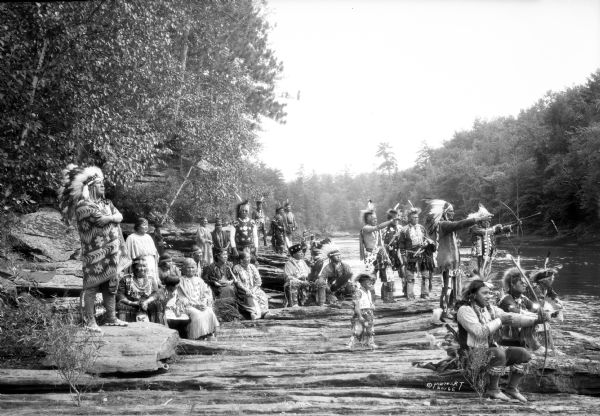 A large group of Native Americans, men, women and children, posing on rocks along the Wisconsin River. They are wearing traditional clothing, and some of them are holding weapons pointed across the river. Native Americans, including Ho-Chunk, Sioux, Kiowa and southwestern Native American tribes, were gathered at Wisconsin Dells for the Stand Rock Indian Ceremonial.