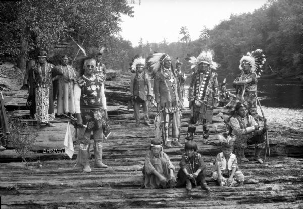Group portrait of Native Americans, including Ho-Chunk, Sioux, Kiowa and southwestern Native American tribes, posing on rocks along the Wisconsin River wearing traditional clothing with beadwork or embroidery. There are three children and two women, all wearing beaded headbands. Nine men are holding either weapons or ceremonial implements, and are wearing feathered headdresses. The tribes were gathered at Wisconsin Dells for the Stand Rock Indian Ceremonial.