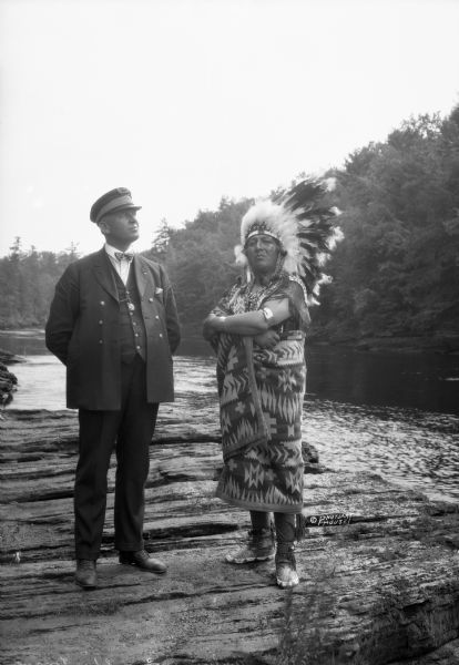 Outdoor portrait of a Native American man with his arms crossed over his chest standing on rocks near the Wisconsin River. He is wearing a headdress, a patterned blanket, a fringed shirt, leather beaded and belled moccasins, and arm and wrist bracelets. Standing next to him on the left is a man wearing a uniform, including a hat that says "Captain" on the front, a bow tie, jacket, and a vest. He is also wearing a medal attached to a chain on his vest.