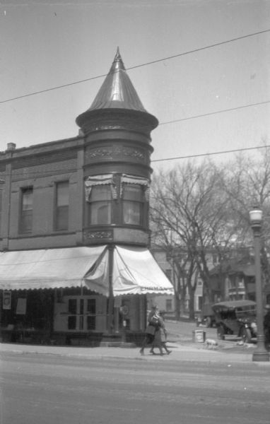 View across street towards Ehrman's Delicatessen, located at 300 State Street, on the corner at Henry Street. Two people are walking on the sidewalk near the entrance to the deli. Under the awning is a large scale, and at the corner near a parked automobile is a dog on the sidewalk.